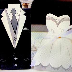 May include: A pair of wedding favour boxes, one shaped like a tuxedo and the other like a wedding dress. The tuxedo box is black with a white shirt and tie. The wedding dress box is white with a grey lace trim and a small gold heart.