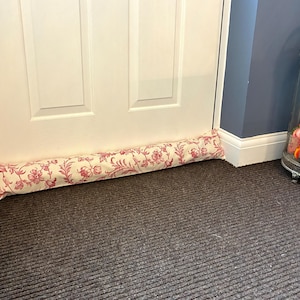 Op de afbeelding: Een stoffen tochtstopper met een crèmekleurige achtergrond en een rood bloemenpatroon. De cilindrische stopper rust tegen een witte deur op een donkerbruin tapijt. Een decoratieve lantaarn staat op de achtergrond.