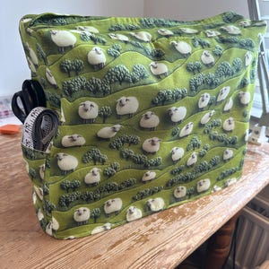 May include: A green fabric sewing machine cover featuring a repeating pattern of white sheep grazing on rolling hills with small trees. The cover includes a side pocket containing a pair of black scissors and a white measuring tape.