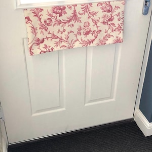 Op de afbeelding: Een witte deur met een stoffen deurstopper met een bloemenpatroon. De deurstopper is roze en wit met een bloemenontwerp. De deurstopper is met een haak aan de bovenkant van de deur bevestigd.