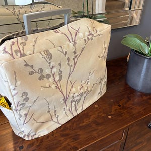 May include: A beige sewing machine cover featuring a floral design in purple and grey. It has a grey handle and a side pocket containing a yellow measuring tape. The cover sits on a wooden surface. The cover's design includes branches and leaves.
