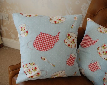May include: Two blue and white throw pillows with a red and white gingham teapot and teacup pattern. The pillows are on a brown leather couch.