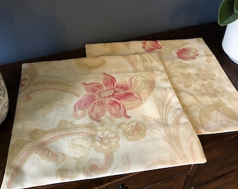 May include: Two cream-colored throw pillows with a floral pattern in shades of pink and brown. The pillows are on a wooden dresser with drawers.