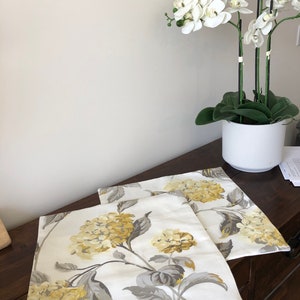 Puede incluir: Dos almohadas blancas con un patrón floral con hortensias amarillas y grises. Las almohadas están sobre una superficie de madera con una planta de orquídea blanca en una maceta blanca en el fondo.