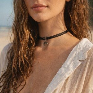 May include: A black choker necklace with a silver crescent moon pendant. The necklace is made of three thin, black cords. The model is wearing a white button-down shirt and a green bikini top.