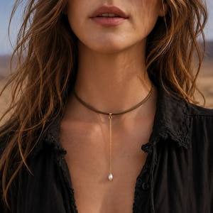 May include: A dark olive green cord choker necklace featuring a thin gold-toned metal chain drop that extends downward from the center. A single small, white, teardrop-shaped pearl is attached to the bottom end of the delicate gold chain.