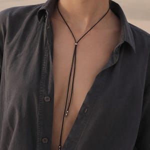 May include: A black lariat necklace with a silver accent. The necklace is worn over a dark grey button-down shirt. The background is a blurred desert landscape.