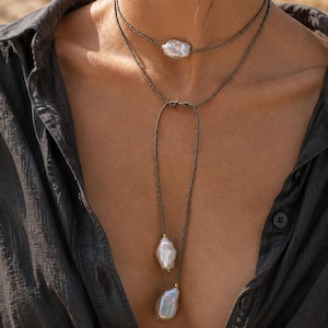 May include: A layered necklace featuring dark metallic beads and three irregular white baroque pearls. The design includes a choker-style strand with one central pearl and a longer lariat-style strand with two dangling pearls at the ends.