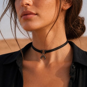May include: A black choker necklace with a crescent moon pendant. The necklace is worn with a black button-down shirt and a black lace bra. The image is taken in natural light.