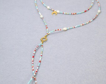 May include: A multi-strand necklace featuring light blue, red, and white seed beads with interspersed white pearls. The design includes a gold-toned sun charm at the center, a small crescent moon charm, and two dangling pearl ends on a gold chain.