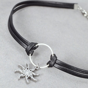 May include: Black leather choker necklace with a silver sun pendant. The necklace features a double-strand design with a silver ring detail and a lobster clasp closure. The sun pendant has a detailed, stylized design.