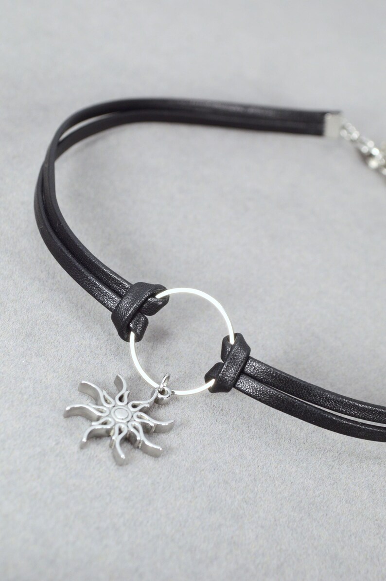 May include: Black leather choker necklace with a silver sun pendant. The necklace features a double-strand design with a silver ring detail and a lobster clasp closure. The sun pendant has a detailed, stylized design.