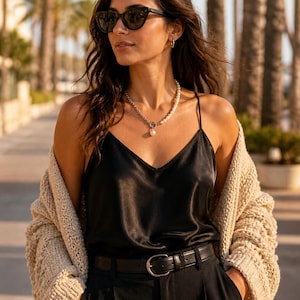 May include: A woman wearing a black silk camisole top, black pants, and a beige knit cardigan. She is also wearing a pearl necklace, sunglasses, and a black belt with a silver buckle. The background features palm trees.