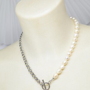 May include: A two-tone necklace featuring a silver chain on one side and a strand of white pearls on the other. The necklace is secured with a silver toggle clasp and a dangling pearl pendant. The jewelry is displayed on a mannequin.