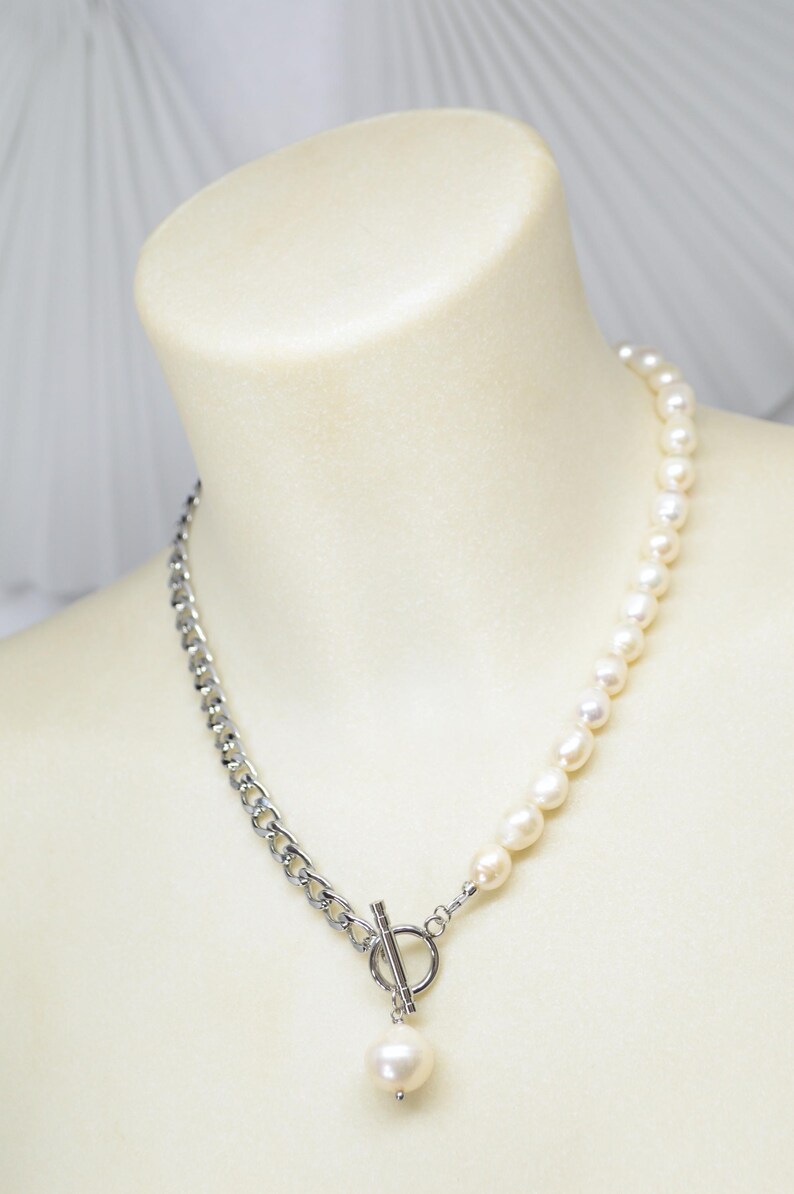 May include: A two-tone necklace featuring a silver chain on one side and a strand of white pearls on the other. The necklace is secured with a silver toggle clasp and a dangling pearl pendant. The jewelry is displayed on a mannequin.