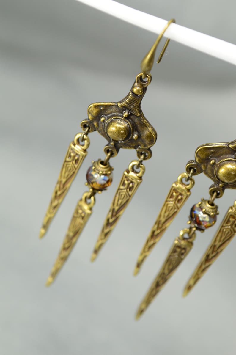 May include: Gold tone dangle earrings with three pointed, ornate drops and a single iridescent bead on each earring.