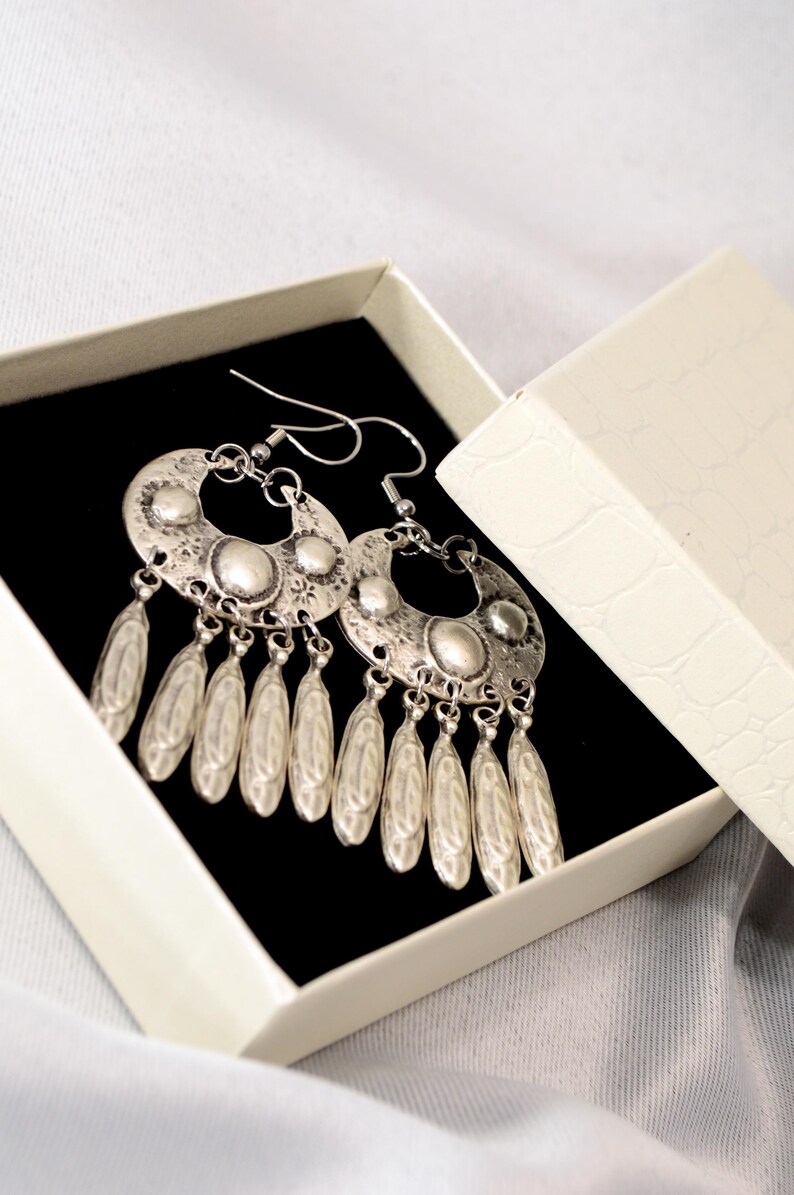 Puede incluir: Pendientes plateados con dise&ntilde;o de media luna y colgantes en forma de l&aacute;grima. Los pendientes se muestran en una caja de joyer&iacute;a blanca abierta con interior negro, sobre un fondo suave y neutro.