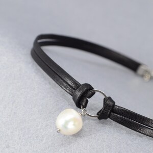 May include: A black leather choker necklace with a large, white pearl pendant. The necklace features a double-strand design with a knotted detail and a silver-colored ring holding the pearl.