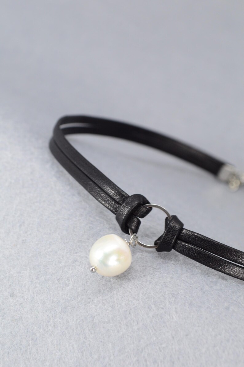 May include: A black leather choker necklace with a large, white pearl pendant. The necklace features a double-strand design with a knotted detail and a silver-colored ring holding the pearl.