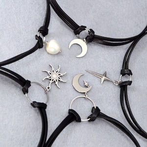 May include: Several black leather choker necklaces with silver-tone charms. Charms include a pearl, crescent moon, sunburst, star, and a crescent moon with a star. The necklaces are displayed on a light gray surface.