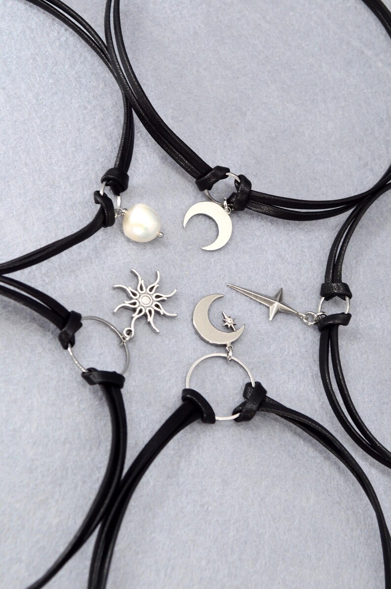 May include: Several black leather choker necklaces with silver-tone charms. Charms include a pearl, crescent moon, sunburst, star, and a crescent moon with a star. The necklaces are displayed on a light gray surface.