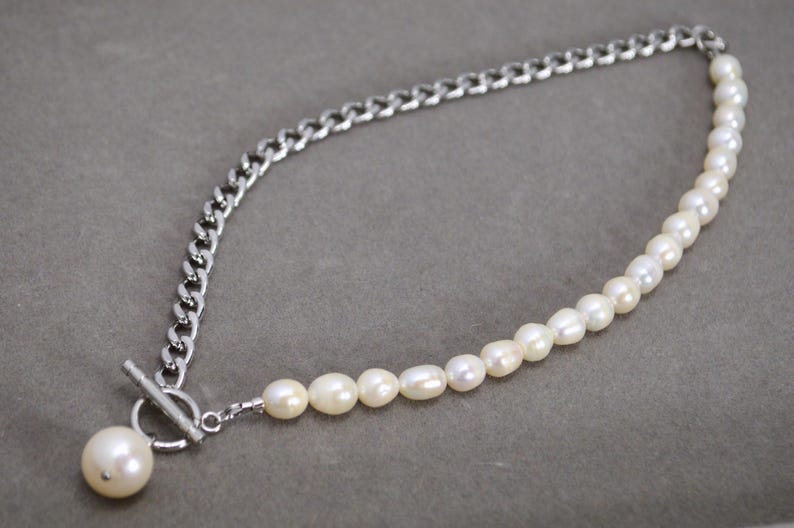 May include: A silver chain necklace with a toggle clasp and a strand of white pearls. The necklace features a chunky chain on one side and a row of pearls on the other, creating a unique design.