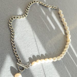 May include: A necklace featuring a silver chain on one side and a strand of pearls on the other. The necklace has a toggle clasp and a single pearl pendant. The jewelry is displayed on a gray surface.