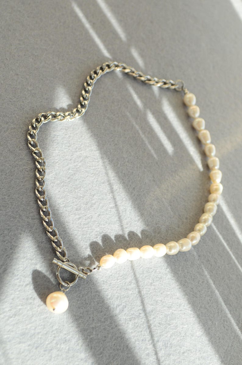 May include: A necklace featuring a silver chain on one side and a strand of pearls on the other. The necklace has a toggle clasp and a single pearl pendant. The jewelry is displayed on a gray surface.