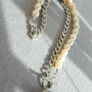 May include: A layered necklace featuring a silver chain and a strand of off-white pearls. The necklace has a toggle clasp and a single pearl pendant. The jewelry is displayed on a gray surface.