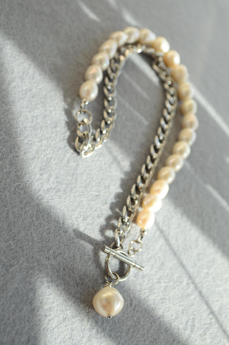 May include: A layered necklace featuring a silver chain and a strand of off-white pearls. The necklace has a toggle clasp and a single pearl pendant. The jewelry is displayed on a gray surface.