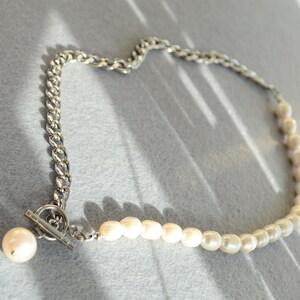 May include: A necklace featuring a silver chain and a strand of white pearls. The chain is a thick, curb style, and the pearls are small and round. The necklace has a toggle clasp and a large pearl pendant.