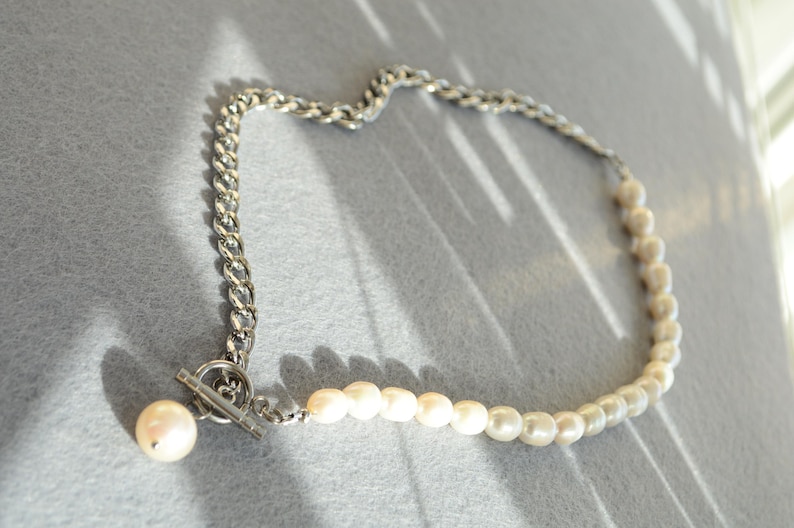 May include: A necklace featuring a silver chain and a strand of white pearls. The chain is a thick, curb style, and the pearls are small and round. The necklace has a toggle clasp and a large pearl pendant.
