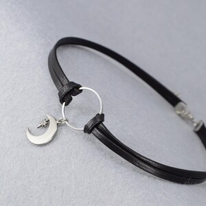 May include: Black leather bracelet featuring a silver-tone crescent moon charm with a star accent. The bracelet has a simple design with a circular connector and a clasp closure. The jewelry is set against a light gray background.