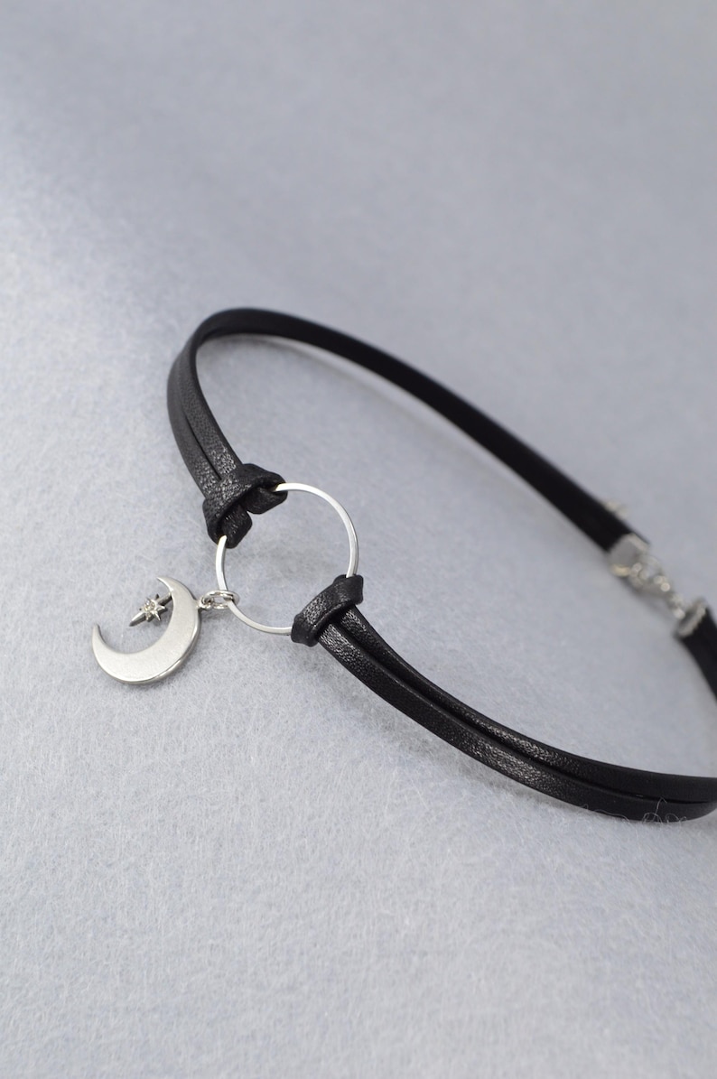 May include: Black leather bracelet featuring a silver-tone crescent moon charm with a star accent. The bracelet has a simple design with a circular connector and a clasp closure. The jewelry is set against a light gray background.