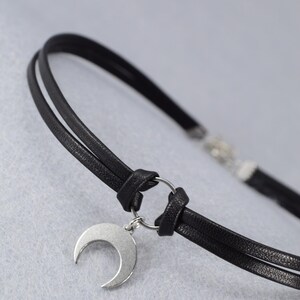 May include: Black choker necklace with a silver crescent moon pendant. The necklace features two thin, black leather cords connected by a silver ring. The crescent moon hangs from the ring, creating a minimalist and celestial design.