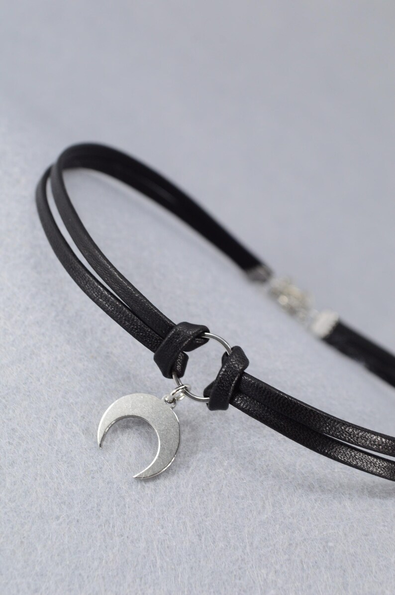 May include: Black choker necklace with a silver crescent moon pendant. The necklace features two thin, black leather cords connected by a silver ring. The crescent moon hangs from the ring, creating a minimalist and celestial design.