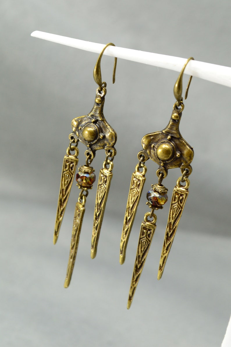 May include: A pair of gold-tone dangle earrings with a geometric design and brown glass beads. The earrings have a long, pointed shape and are suspended from a hook.