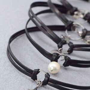 May include: Black leather chokers with silver-tone accents. One choker features a pearl, and another has a cross pendant. The chokers are displayed on a light gray surface.