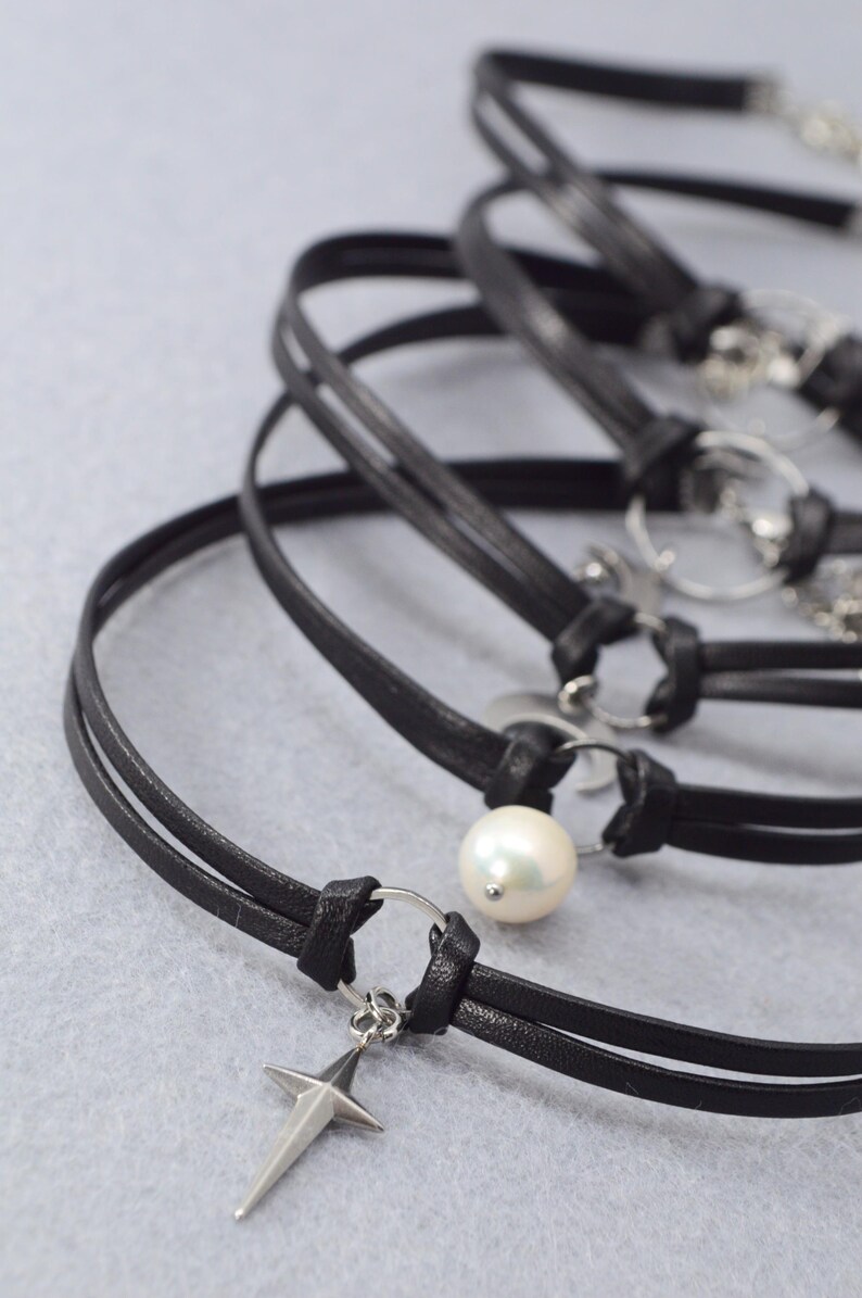May include: Black leather chokers with silver-tone accents. One choker features a pearl, and another has a cross pendant. The chokers are displayed on a light gray surface.