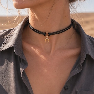 Op de afbeelding: Een zwarte choker-halsketting met dubbele koorden en een kleine goudkleurige halvemaanvormige hanger die aan een centrale goudkleurige ring hangt. De ketting zit om de hals en rust op een grijs overhemd met kraag en knoopsluiting.