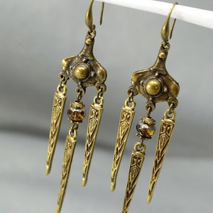 May include: A pair of gold-tone dangle earrings with three teardrop-shaped pendants each. The pendants have an intricate geometric design and are accented with a small amber-coloured glass bead.
