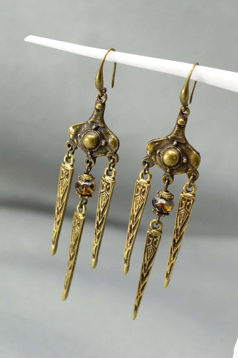 May include: A pair of gold-tone dangle earrings with three teardrop-shaped pendants each. The pendants have an intricate geometric design and are accented with a small amber-coloured glass bead.