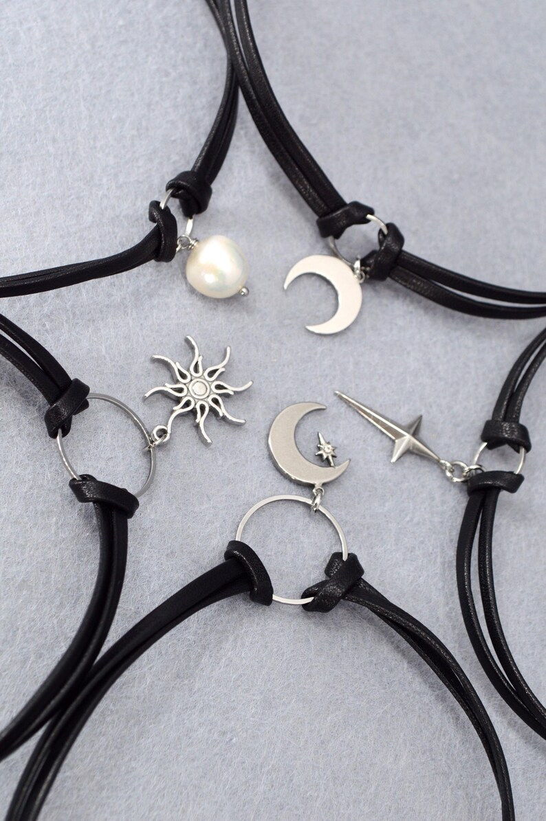 May include: Black leather choker necklaces with silver-tone celestial charms. The charms include a pearl, crescent moons, a sun, and a star. The necklaces are displayed on a gray surface.