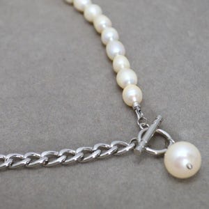 May include: A necklace featuring a strand of white pearls connected to a silver chain. The necklace has a toggle clasp and a large pearl pendant. The jewelry is set against a gray background.