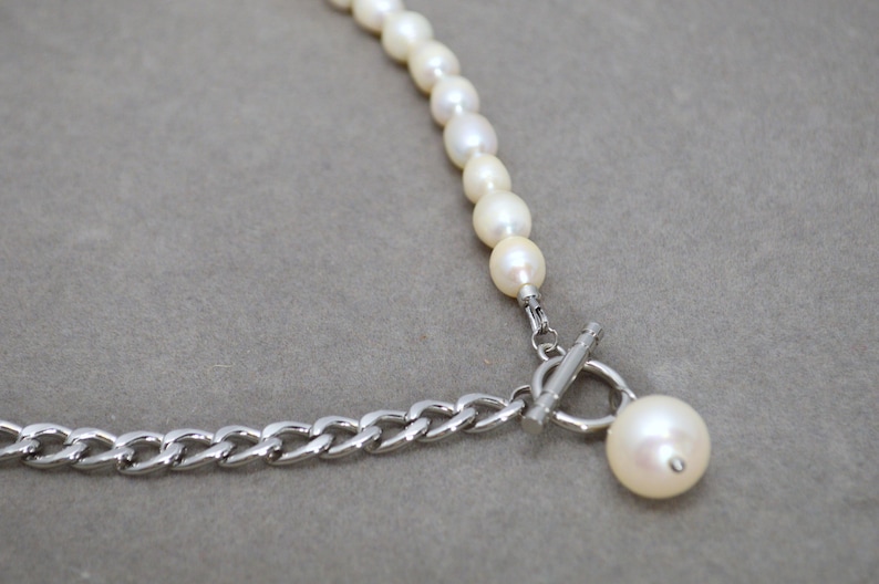 May include: A necklace featuring a strand of white pearls connected to a silver chain. The necklace has a toggle clasp and a large pearl pendant. The jewelry is set against a gray background.