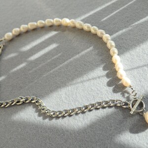 May include: A necklace featuring a combination of pearls and a silver chain. The necklace has a strand of small, off-white pearls on one side and a silver chain on the other. A silver toggle clasp and a single pearl pendant complete the design.