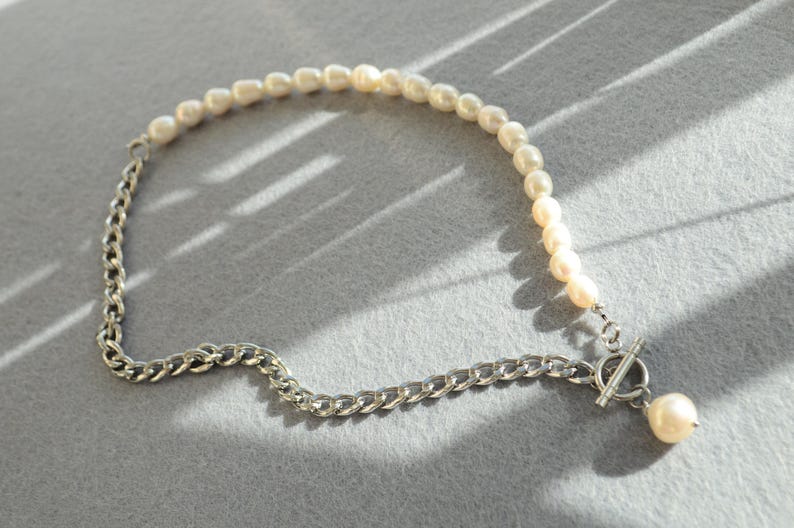 May include: A necklace featuring a combination of pearls and a silver chain. The necklace has a strand of small, off-white pearls on one side and a silver chain on the other. A silver toggle clasp and a single pearl pendant complete the design.