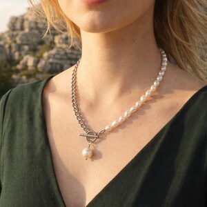 May include: A silver chain and pearl necklace. The necklace features a silver chain on one side and a strand of pearls on the other. A silver toggle clasp connects the chain and pearl strand. A single pearl pendant hangs from the clasp.