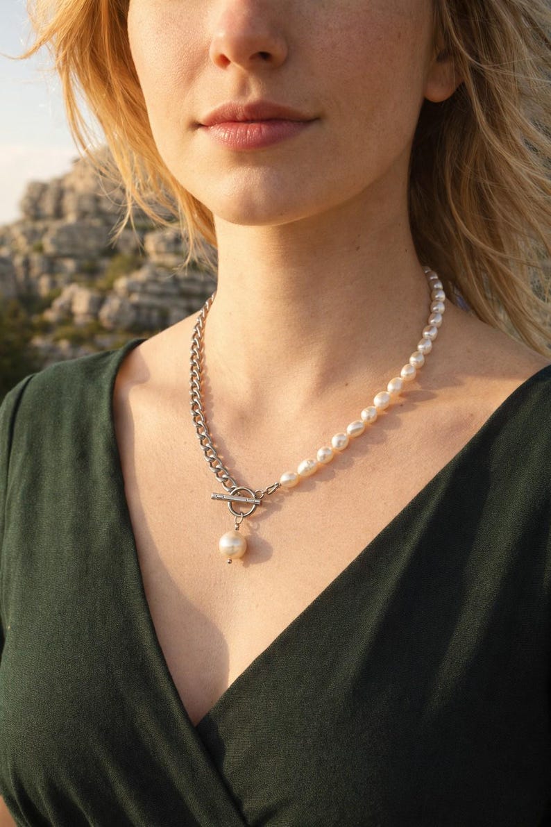 May include: A silver chain and pearl necklace. The necklace features a silver chain on one side and a strand of pearls on the other. A silver toggle clasp connects the chain and pearl strand. A single pearl pendant hangs from the clasp.