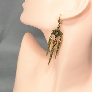May include: Gold tone dangle earrings with a geometric design and a single red bead. The earrings are on a mannequin's ear.
